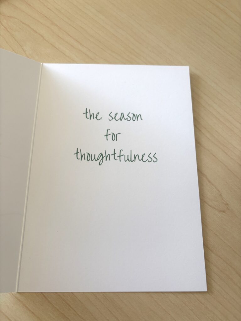 An open holiday card rests on a light wood surface. The inside right panel is white with the message "the season for thoughtfulness" centered in dark green, handwritten-style lettering, leaving space below for a personal note.